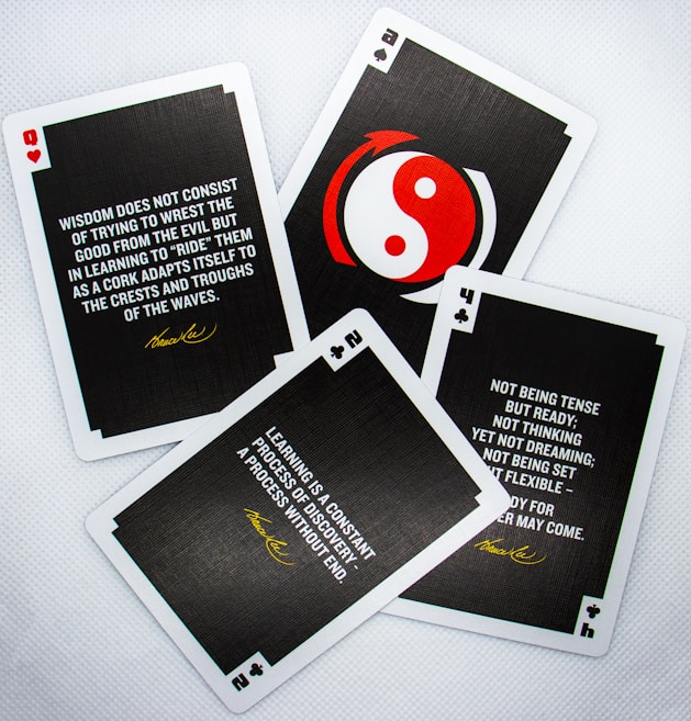 Four playing cards are arranged on a white surface. Each card contains a motivational quote attributed to Bruce Lee. The cards display different numbers and suits: hearts, clubs, and spades. One card features a red and white yin-yang symbol in the center.