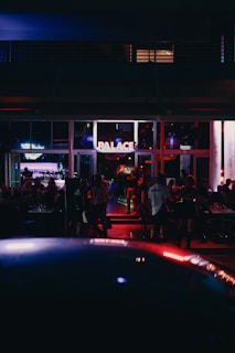 A dimly lit venue with people socializing inside. The area includes tables and chairs and is illuminated by various colorful lights. A sign with the word 'PALACE' is prominently displayed above the entrance. The atmosphere suggests nighttime with indoor and outdoor elements visible.