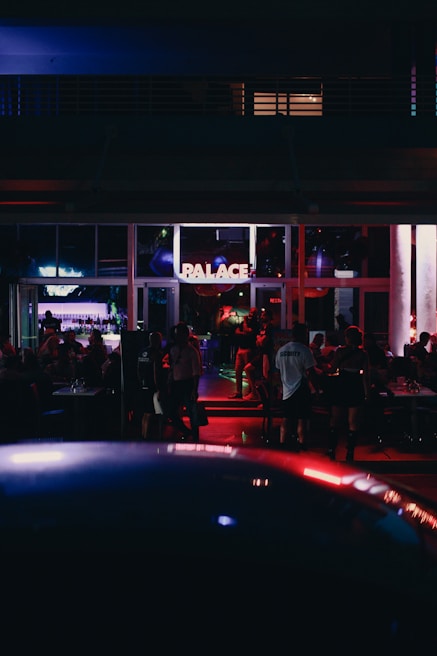 A dimly lit venue with people socializing inside. The area includes tables and chairs and is illuminated by various colorful lights. A sign with the word 'PALACE' is prominently displayed above the entrance. The atmosphere suggests nighttime with indoor and outdoor elements visible.