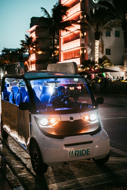 A small electric vehicle with bright headlights is parked on a city street at night. The street is illuminated by surrounding neon lights reflected from a nearby building, creating a lively and vibrant atmosphere. Palm trees are visible, suggesting a warm, urban environment.
