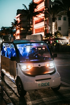 A small electric vehicle with bright headlights is parked on a city street at night. The street is illuminated by surrounding neon lights reflected from a nearby building, creating a lively and vibrant atmosphere. Palm trees are visible, suggesting a warm, urban environment.