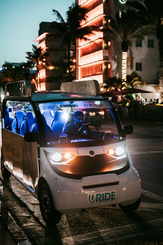 A small electric vehicle with bright headlights is parked on a city street at night. The street is illuminated by surrounding neon lights reflected from a nearby building, creating a lively and vibrant atmosphere. Palm trees are visible, suggesting a warm, urban environment.