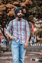 A person with a turban and mustache, wearing a plaid shirt and jeans, is standing outdoors. The background features trees with colorful foliage and people in the distance.