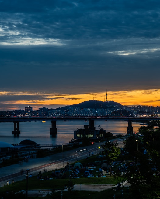 A vibrant cityscape at dusk with a river reflecting the lights of a bridge and an urban skyline in the background. A prominent tower is visible on a hill, silhouetted against a sunset sky. The foreground features a park with people gathering and paths illuminated by streetlights.
