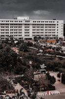 A large multi-story academic building labeled 'Academic Block - 7' stands prominently. The structure is surrounded by lush greenery and well-maintained gardens. There are several yellow buses parked in front of the building, along with pathways and a few people scattered around the campus grounds.