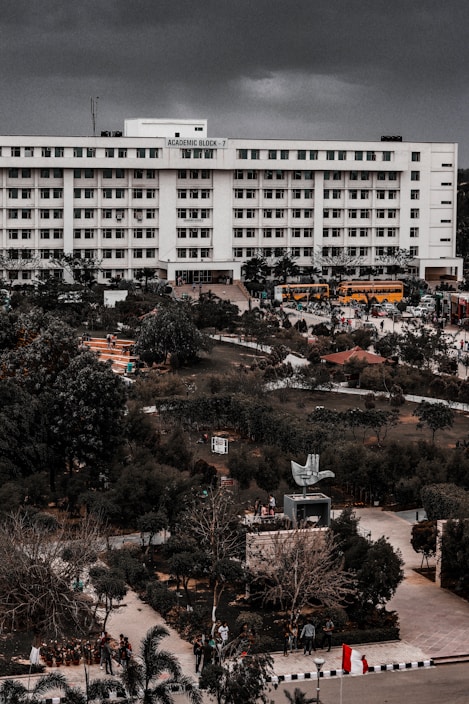 A large multi-story academic building labeled 'Academic Block - 7' stands prominently. The structure is surrounded by lush greenery and well-maintained gardens. There are several yellow buses parked in front of the building, along with pathways and a few people scattered around the campus grounds.