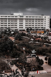 A large multi-story academic building labeled 'Academic Block - 7' stands prominently. The structure is surrounded by lush greenery and well-maintained gardens. There are several yellow buses parked in front of the building, along with pathways and a few people scattered around the campus grounds.