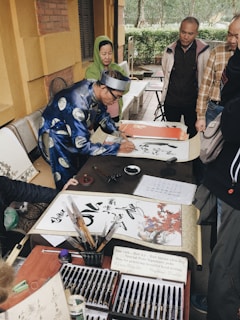 A person dressed in traditional attire is engaged in calligraphy with several people watching. The setting is outdoors, with stone walls and greenery visible in the background. Various calligraphy tools and papers are spread across the table.