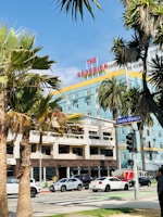 Palm trees frame a colorful hotel with a retro sign reading 'The Georgian'. Below, cars move along a busy street featuring a traffic light and a sidewalk with pedestrians. The sky is clear and blue.
