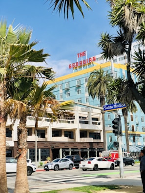 Palm trees frame a colorful hotel with a retro sign reading 'The Georgian'. Below, cars move along a busy street featuring a traffic light and a sidewalk with pedestrians. The sky is clear and blue.