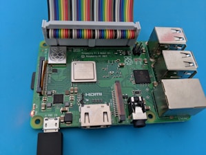 A circuit board with multiple electronic components, featuring ports for USB, HDMI, Ethernet, and power. A colorful ribbon cable is connected to its pins. It's laid on a blue surface.