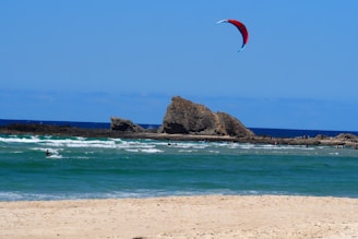 A sandy beach stretches towards the turquoise ocean waves where a person is kite surfing. A large rocky formation is visible in the background, and several people can be seen playing in the shallow waters. A vibrant red and blue kite is flying high in the sky against a clear, sunny backdrop.