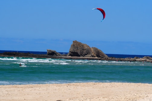 A sandy beach stretches towards the turquoise ocean waves where a person is kite surfing. A large rocky formation is visible in the background, and several people can be seen playing in the shallow waters. A vibrant red and blue kite is flying high in the sky against a clear, sunny backdrop.
