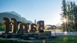 Large metallic letters spelling out a name are prominently placed on rocks in the foreground. In the background, the sunlight filters through tall pine trees, with mountains visible under a clear blue sky. The scene has a calm, serene morning vibe, with a few buildings visible along a paved pathway.