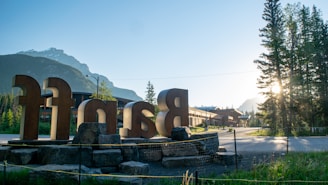 Large metallic letters spelling out a name are prominently placed on rocks in the foreground. In the background, the sunlight filters through tall pine trees, with mountains visible under a clear blue sky. The scene has a calm, serene morning vibe, with a few buildings visible along a paved pathway.