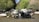 A motorcycle is parked in a shallow stream surrounded by rocks and lush greenery, including trees and bushes. The motorcycle is loaded with gear, highlighting a sense of adventure and exploration.