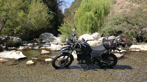 An adventure bike crossing a shallow river surrounded by lush forest.