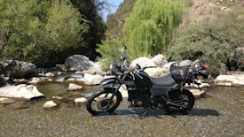 A motorcycle is parked in a shallow stream surrounded by rocks and lush greenery, including trees and bushes. The motorcycle is loaded with gear, highlighting a sense of adventure and exploration.