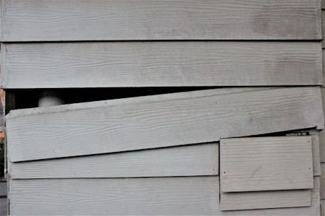 The image displays a section of overlapping wooden siding planks. The planks have a horizontal orientation and feature a weathered, aged texture with visible wood grain. There is a noticeable broken or misaligned plank in the middle, creating an uneven appearance.