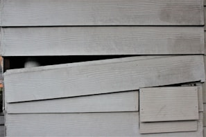 The image displays a section of overlapping wooden siding planks. The planks have a horizontal orientation and feature a weathered, aged texture with visible wood grain. There is a noticeable broken or misaligned plank in the middle, creating an uneven appearance.