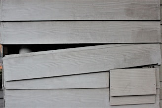 The image displays a section of overlapping wooden siding planks. The planks have a horizontal orientation and feature a weathered, aged texture with visible wood grain. There is a noticeable broken or misaligned plank in the middle, creating an uneven appearance.