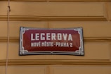 A street sign with a red background and white lettering is mounted on a yellow textured wall. The text on the sign reads 'LEGEROVA' with 'NOVÉ MĚSTO-PRAHA 2' beneath it. The sign features a decorative blue and white border.