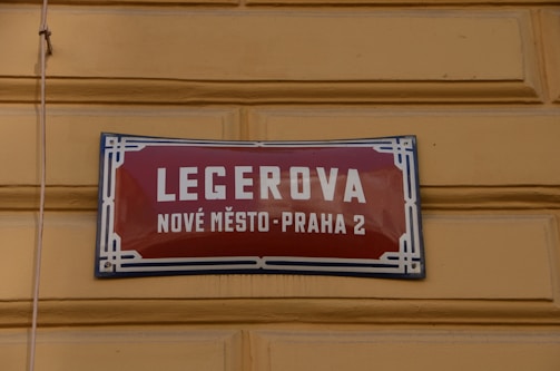 A street sign with a red background and white lettering is mounted on a yellow textured wall. The text on the sign reads 'LEGEROVA' with 'NOVÉ MĚSTO-PRAHA 2' beneath it. The sign features a decorative blue and white border.