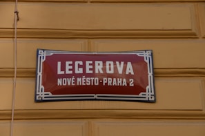 A street sign with a red background and white lettering is mounted on a yellow textured wall. The text on the sign reads 'LEGEROVA' with 'NOVÉ MĚSTO-PRAHA 2' beneath it. The sign features a decorative blue and white border.