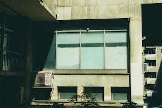 A large window on an industrial-looking building set against a backdrop of concrete walls. The window has a couple of glass panels, partially covered or tinted, with some visible reflections. There's a metallic structure attached to the wall beside the window. Shadows and light patterns create contrast on the building's facade.