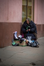 An older woman wearing a headscarf and warm clothing is seated against a building wall. In front of her, there are various small items for sale, including shoes and socks, placed on a cardboard box and in plastic bags.