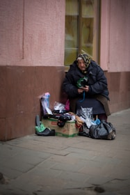 An older woman wearing a headscarf and warm clothing is seated against a building wall. In front of her, there are various small items for sale, including shoes and socks, placed on a cardboard box and in plastic bags.