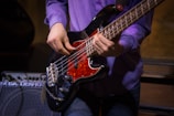 A person is playing a black electric bass guitar with a red pickguard. The individual's hands are prominently positioned on the strings, suggesting they are in the midst of a performance. The person is wearing a purple shirt and blue jeans. In the background, there is an amplifier with visible text on it.