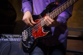 A person is playing a black electric bass guitar with a red pickguard. The individual's hands are prominently positioned on the strings, suggesting they are in the midst of a performance. The person is wearing a purple shirt and blue jeans. In the background, there is an amplifier with visible text on it.