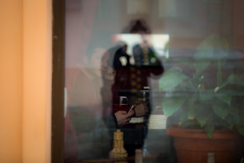 A reflective window scene showing a person inside a dimly lit room holding a phone in one hand and resting their head on the other. The person appears contemplative and seems to be seated at a table with drinks. The window reflection also shows a large green potted plant and another individual in the background.