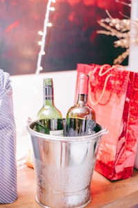 Two bottles of wine are placed inside a metallic ice bucket. The bucket is on a wooden surface with water droplets on its exterior, indicating it is cold. In the background, there are red and white gift bags and strings of soft glowing lights, contributing to a festive atmosphere.