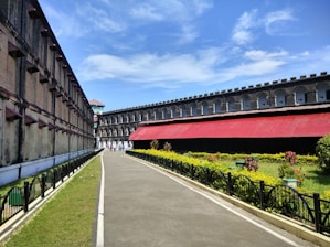 A pathway runs between two long, historic buildings made from stone and brick. One building has a red roof and the area is surrounded by manicured grass and a garden with flowers and shrubs. The sky is clear and blue, adding to the bright, sunny atmosphere.