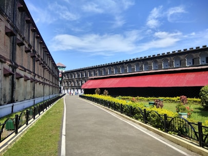 A pathway runs between two long, historic buildings made from stone and brick. One building has a red roof and the area is surrounded by manicured grass and a garden with flowers and shrubs. The sky is clear and blue, adding to the bright, sunny atmosphere.