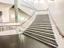 A wide staircase with sleek, modern design characterized by white walls and railings. The steps are black with geometric patterns on the risers. Soft lighting enhances the clean aesthetic, and a decorative blue vertical element adds contrast.