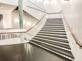 A wide staircase with sleek, modern design characterized by white walls and railings. The steps are black with geometric patterns on the risers. Soft lighting enhances the clean aesthetic, and a decorative blue vertical element adds contrast.