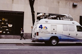 A news van parked on a city street adjacent to a building with a large window display. The van, marked with NBC New York branding and logos, features equipment on the roof likely for broadcasting. Two individuals walk on the sidewalk, one holding a coffee cup. The street and building have a muted, urban atmosphere.