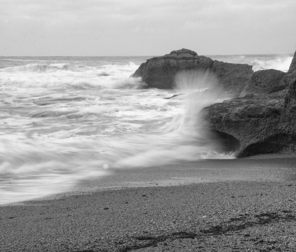 Fotografia in scala di grigi dell'onda del mare che schizza sulla roccia