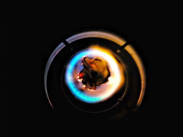 A close-up of a gas stove burner with a blue and yellow flame surrounding a dark central structure, set against a black background.