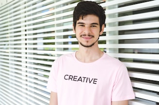 A young man with dark hair and a beard is standing in front of a wall with horizontal white slats. He is wearing a light pink t-shirt with the word 'CREATIVE' printed in bold, black letters. The lighting is bright and the background has some greenery visible through the slats.