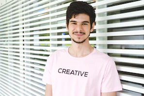 A young man with dark hair and a beard is standing in front of a wall with horizontal white slats. He is wearing a light pink t-shirt with the word 'CREATIVE' printed in bold, black letters. The lighting is bright and the background has some greenery visible through the slats.