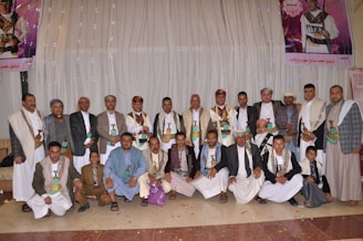 A group of men and boys dressed in traditional attire, including long robes and sashes, are gathered in a row indoors. Many are wearing head coverings, and some have ceremonial daggers (jambiyas) in their belts. In the background, there are decorative banners with images positioned on the walls.