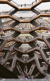 A large, intricate honeycomb-like structure with multiple levels and staircases. People are walking on the various levels, and the background includes a river and urban buildings.