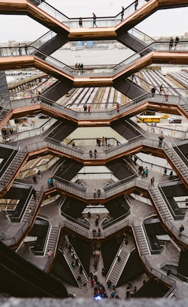 A large, intricate honeycomb-like structure with multiple levels and staircases. People are walking on the various levels, and the background includes a river and urban buildings.