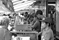 A busy outdoor food stall features a line of workers in uniforms assembling and serving food to a crowd of customers. Overhead lights illuminate the scene, creating a bustling atmosphere.