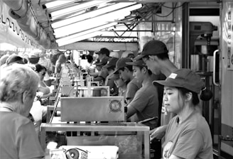 A busy outdoor food stall features a line of workers in uniforms assembling and serving food to a crowd of customers. Overhead lights illuminate the scene, creating a bustling atmosphere.