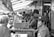 A busy outdoor food stall features a line of workers in uniforms assembling and serving food to a crowd of customers. Overhead lights illuminate the scene, creating a bustling atmosphere.